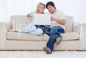 A smiling couple are sitting on a couch looking at a laptop