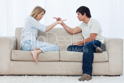 A couple sitting on a couch are pointing at each other