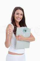 Smiling brunette putting her thumbs up while holding weighing sc