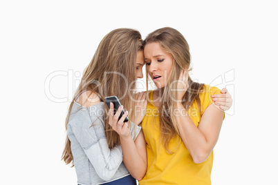 Upset young woman looking her cellphone consolded by her friend