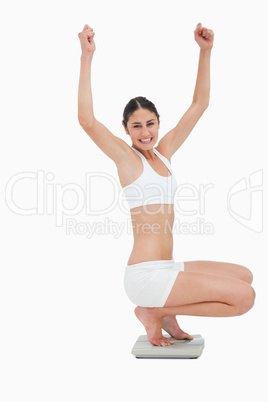 Slim young woman sitting on a scales while raising her arms