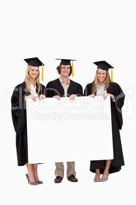 Three students in graduate robe holding a blank sign