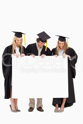 Three smiling students in graduate robe holding a blank sign