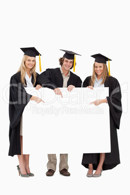 Three students in graduate robe holding and pointing a blank sig