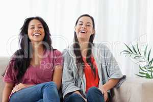 Two women sit beside on another on the couch while laughing