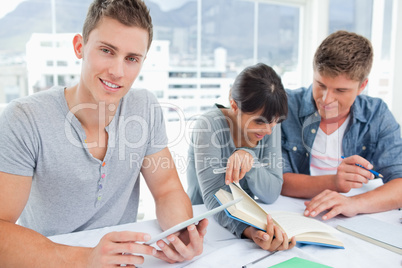 Three smiling students sitting and doing work as one looks at th