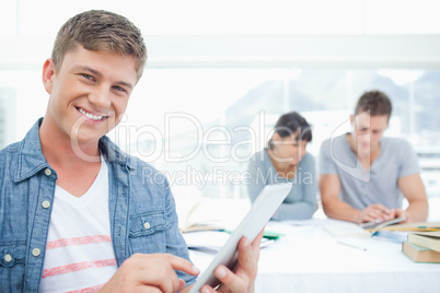 A smiling student using his tablet with his friends in the backg