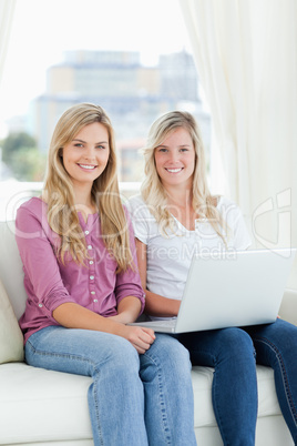 Two sisters sit together and smile with a laptop while looking a