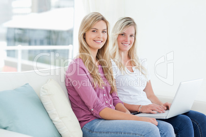 Smiling sisters hold a laptop as they look sideways into the cam