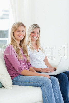 Two sisters sit on the couch with a laptop as they smile at the