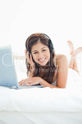 Woman with headphones and a laptop lying on bed, looking forward