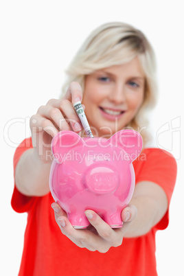 Young blonde woman putting notes into a pink piggy bank