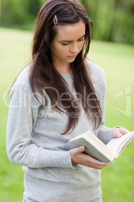 Serious young woman reading a book while standing up in a park