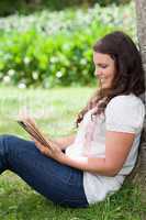 Young smiling woman reading a book while leaning against a tree