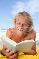 Smiling blonde man reading a book while lying on the beach
