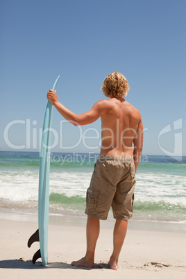 Young blonde man holding his surfboard in front of the ocean