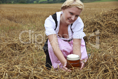Frau im Dirndl kniet mit Bierglas in einem Feld