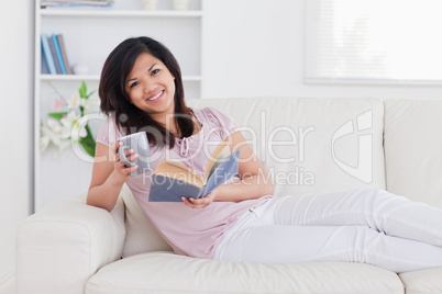 Smiling woman holding a book and a mug while lying on a couch Smiling woman holding a book and a mug while lying on a couch