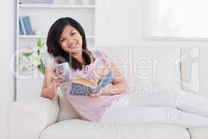 Smiling woman holding a book and a mug while lying on a couch