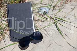 Schwarze Sonnenbrille liegt vor einem Buch am Strand