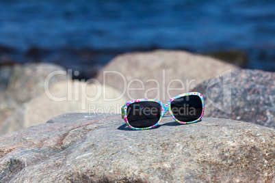 Sonnenbrille mit buntem Gestell liegt am Strand auf einem Felsen