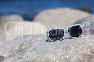 Sonnenbrille mit buntem Gestell liegt am Strand auf einem Felsen