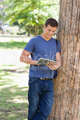 Male student leaning against a tree while using a touch pad