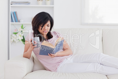 Woman lying on a sofa while holding a mug and a book