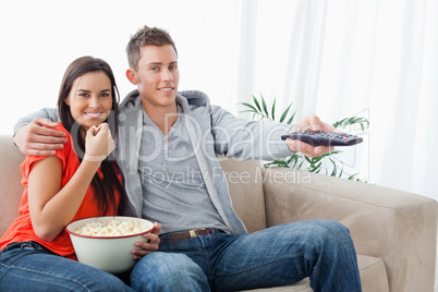 A couple eating popcorn and using the tv remote while looking at