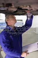 Mechanic touching the below of a car while holding a clipboard