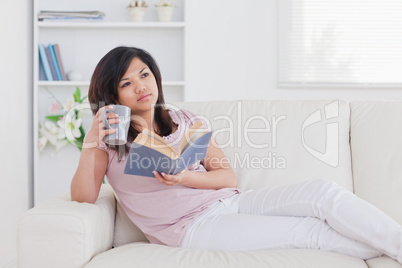 Woman lying on a couch while holding a book and a mug