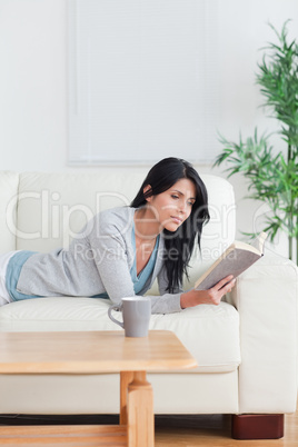 Woman reading a book with a mug on a table while resting on a co