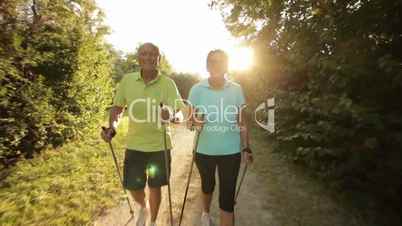 senior nordic walking couple smiling into cam following nordic walkers in park