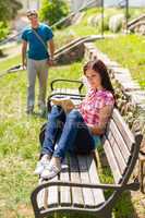 Woman reading book on bench man coming
