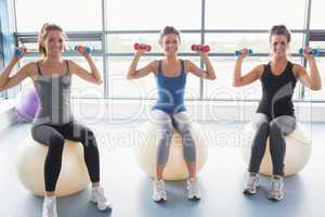 Three smiling women sitting on exercise balls and lifting weight