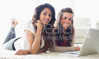 Two smiling women typing on a computer while lying on the floor