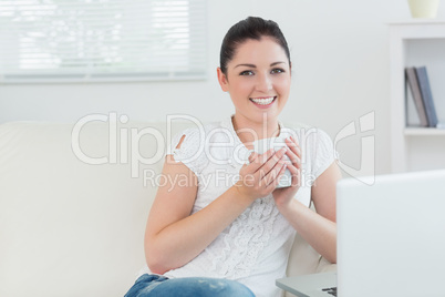 Woman on a couch and holding a cup while using a laptop