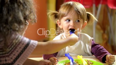 Happy little girl feeding her friend with lasagna in kindergarten