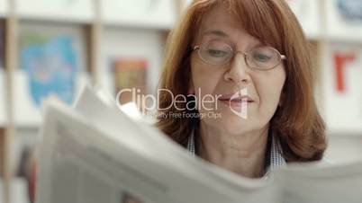 Elderly woman with glasses reading newspaper in library