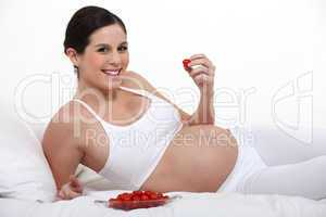 A pregnant woman eating strawberries on her bed.
