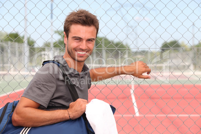 Man with sports bag stood by clay tennis court