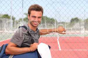 Man with sports bag stood by clay tennis court