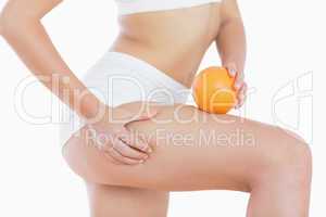 Woman squeezes cellulite skin on thigh as she holds orange