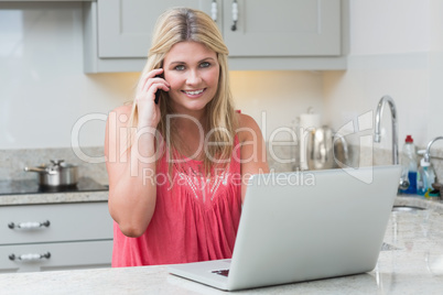 Happy woman using laptop while on call in the kitchen