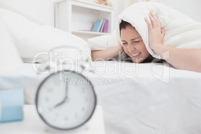 Woman covers ears with sheet as alarm clock rings