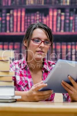 College student using digital tablet with stack of books at libr