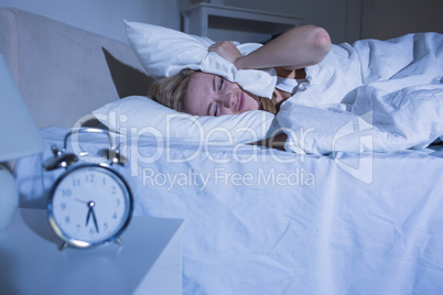 Woman covering ears with pillow as alarm clock rings