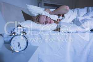 Woman covering ears with pillow as alarm clock rings