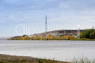 High voltage line and electricity pylon on coastline of river.