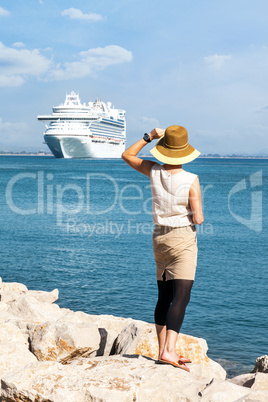Woman with hat stands and looks at the sea, a cruise ship